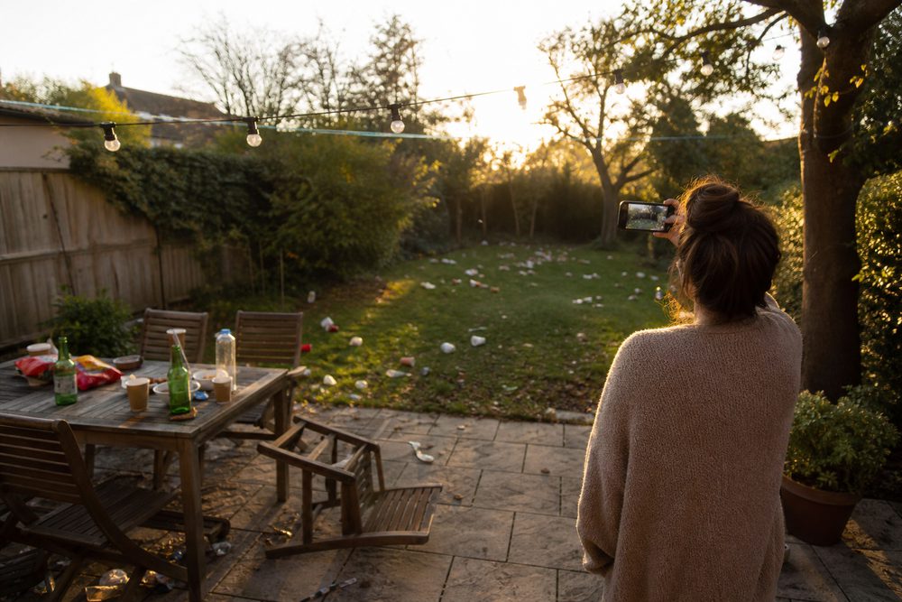 Someone recording a back garden on a phone the morning after a party, cups and bottles scattered across the patio and lawn